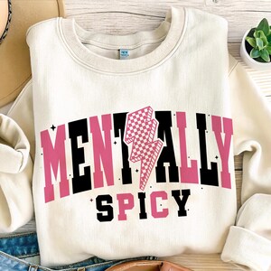 May include: Cream-colored sweatshirt with the words "MENTALLY SPICY" in black and pink block letters. A pink and white checkered lightning bolt graphic is centered above the word "SPICY". The sweatshirt is folded.