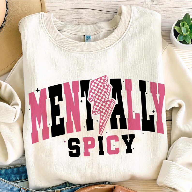 May include: Cream-colored sweatshirt with the words "MENTALLY SPICY" in black and pink block letters. A pink and white checkered lightning bolt graphic is centered above the word "SPICY". The sweatshirt is folded.