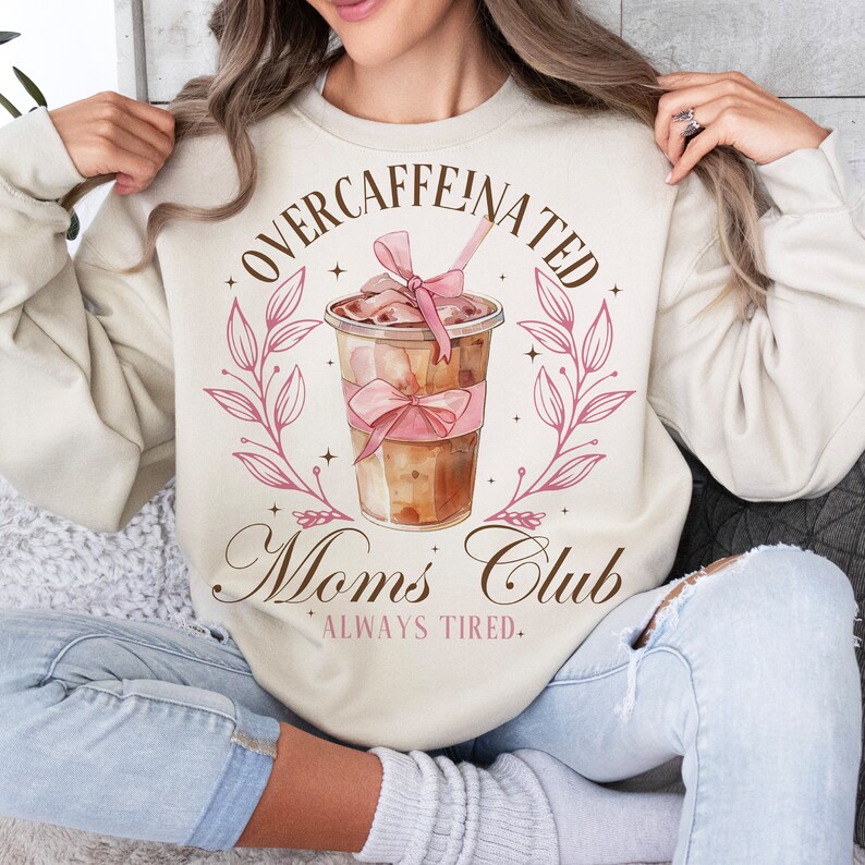 May include: A beige sweatshirt with the text "Overcaffeinated Moms Club Always Tired" and a pink and brown illustration of a coffee drink with a pink bow.
