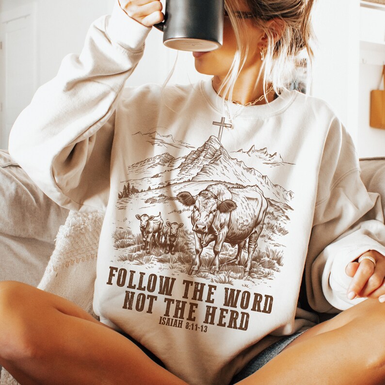 May include: A white sweatshirt with a brown graphic of a cow and two calves in a field with mountains and a cross in the background. The text "FOLLOW THE WORD NOT THE HERD" and "ISAIAH 8:11-13" is printed on the sweatshirt.