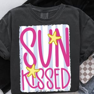 Puede incluir: Camiseta gris oscuro con un dise&ntilde;o gr&aacute;fico. El dise&ntilde;o presenta las palabras "SUN KISSED" en rosa brillante, con acentos de estrellas de mar amarillas, sobre un fondo a rayas azules y blancas.