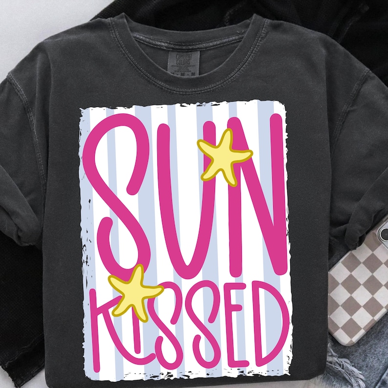 Puede incluir: Camiseta gris oscuro con un dise&ntilde;o gr&aacute;fico. El dise&ntilde;o presenta las palabras "SUN KISSED" en rosa brillante, con acentos de estrellas de mar amarillas, sobre un fondo a rayas azules y blancas.