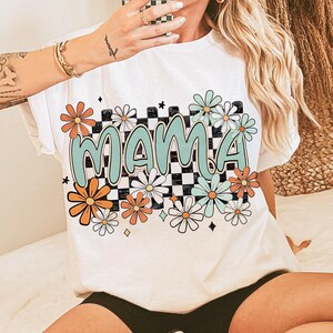 Puede incluir: Camiseta blanca con un patr&oacute;n a cuadros y la palabra "MAMA" en letras verdes. El dise&ntilde;o presenta margaritas coloridas y estrellas.