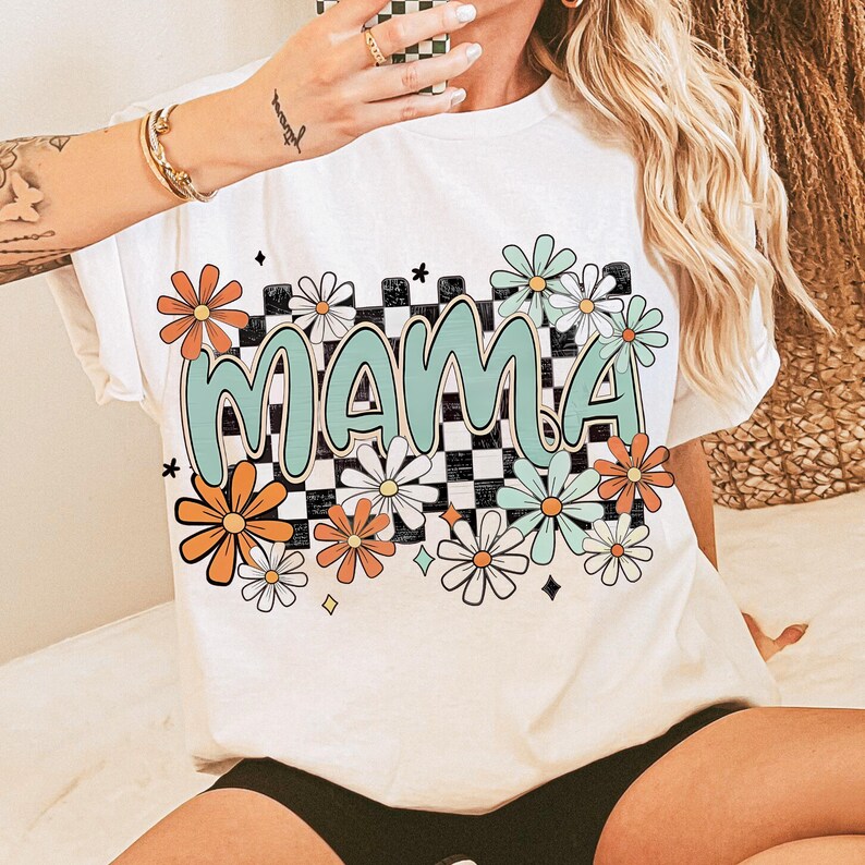 Puede incluir: Camiseta blanca con un patr&oacute;n a cuadros y la palabra "MAMA" en letras verdes. El dise&ntilde;o presenta margaritas coloridas y estrellas.
