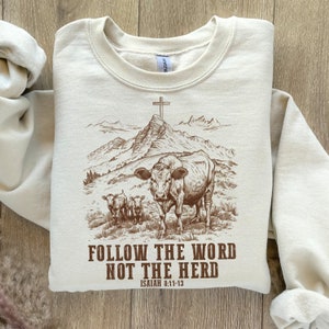 May include: A beige sweatshirt with a brown graphic of a cow and two calves walking towards a mountain with a cross on top. The text "FOLLOW THE WORD NOT THE HERD" is printed below the graphic, with "ISAIAH 8:11-13" printed below the text.