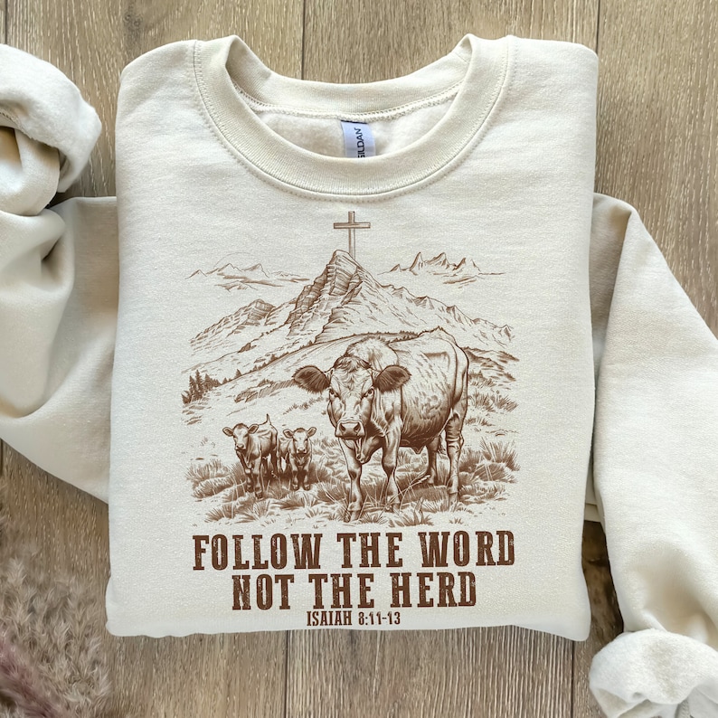 May include: A beige sweatshirt with a brown graphic of a cow and two calves walking towards a mountain with a cross on top. The text "FOLLOW THE WORD NOT THE HERD" is printed below the graphic, with "ISAIAH 8:11-13" printed below the text.