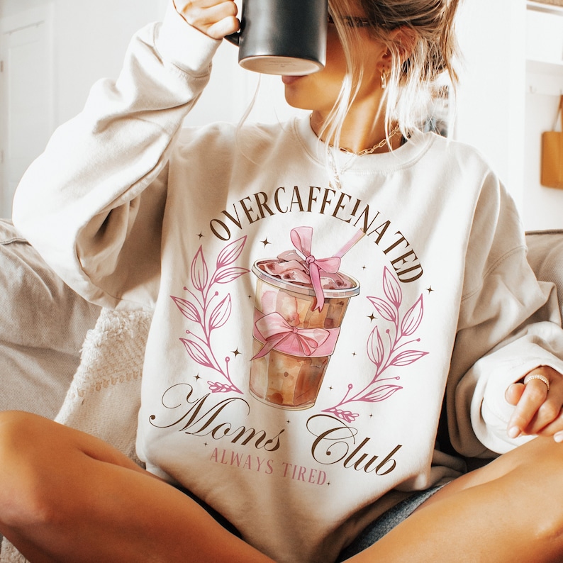 May include: White sweatshirt with a brown and pink graphic of a coffee cup with a pink bow and the text "Overcaffeinated Moms Club Always Tired."