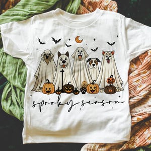Pu&ograve; includere: T-shirt bianca con un design di Halloween. Il design include cinque cani vestiti da fantasmi, zucche, pipistrelli, una luna crescente e la scritta "spooky season". Una maglietta divertente e festosa.