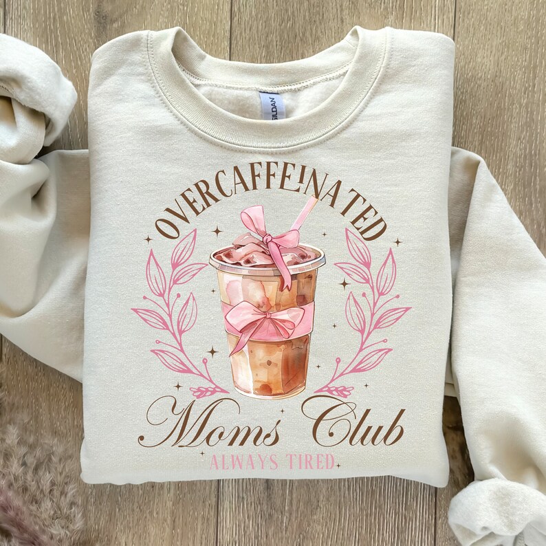 May include: Beige sweatshirt with a brown graphic of a coffee cup with a pink bow and the text "Overcaffeinated Moms Club Always Tired."