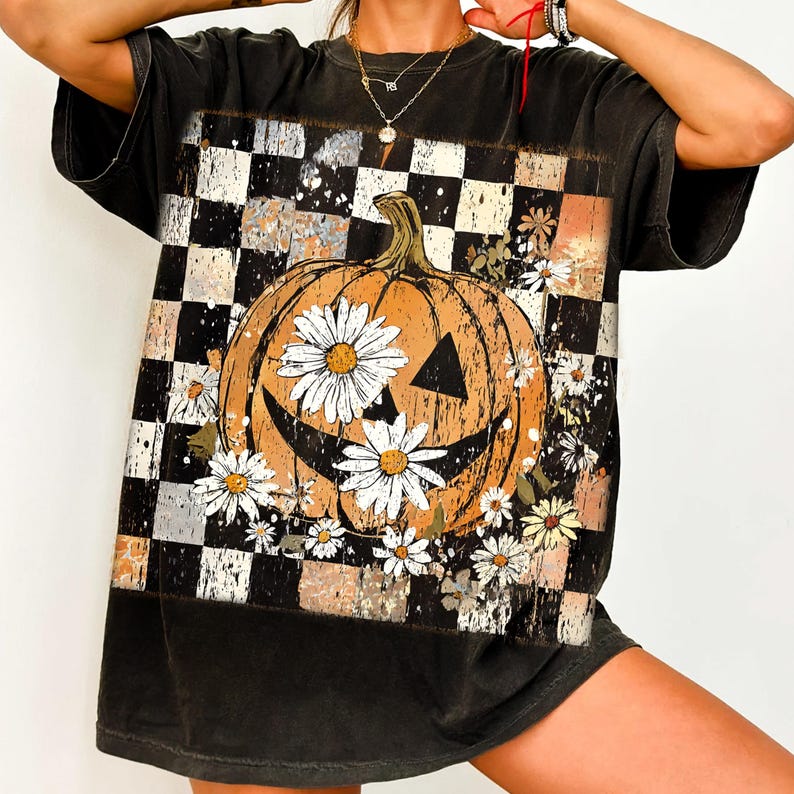 May include: Black t-shirt featuring a smiling jack-o'-lantern design with daisy flowers. The pumpkin is set against a black and white checkered background with orange and brown accents. The shirt has short sleeves and a relaxed fit.