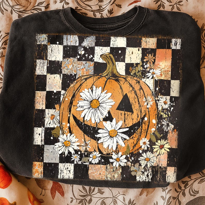 May include: Black crewneck sweatshirt featuring a distressed checkerboard pattern and a smiling orange pumpkin decorated with white daisies. The pumpkin has a carved triangle nose and eyes. A fall-themed design.