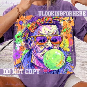 May include: A purple t-shirt featuring a colorful graphic of a character wearing a patterned headscarf and orange-rimmed sunglasses, blowing a large green bubble. The design includes vibrant paint splatters, stars, and the text ULOOKINGFORHERE.