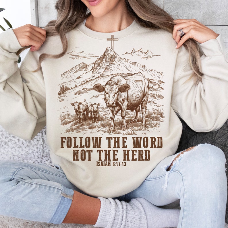 May include: A beige sweatshirt with a brown illustration of a cow and two calves in a field with mountains in the background. The text "FOLLOW THE WORD NOT THE HERD" is printed in brown below the illustration. The text "ISAIAH 8:11-13" is printed in brown below the text.