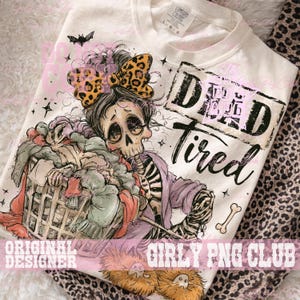 May include: A cream-colored t-shirt featuring a graphic of a skeleton with a leopard print bow, holding a laundry basket filled with clothes. The text reads "DEAD Tired" in a distressed black font, accompanied by small star and bone illustrations.