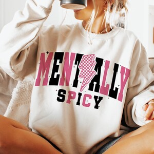 May include: Cream-colored sweatshirt with the words "MENTALLY SPICY" in black and pink block letters. A pink and white checkered lightning bolt graphic is centered above the word "SPICY".