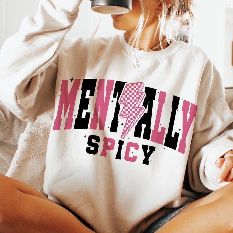 May include: Cream-colored sweatshirt with the words "MENTALLY SPICY" in black and pink block letters. A pink and white checkered lightning bolt graphic is centered above the word "SPICY".