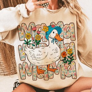 May include: A beige sweatshirt with a white duck wearing a blue hat and a floral pattern. The text "Mama" is repeated three times in a floral design.