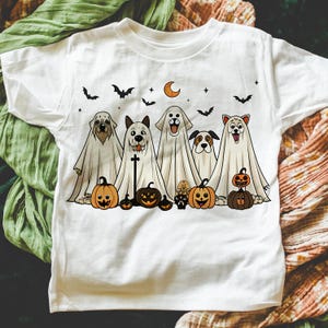 Pu&ograve; includere: T-shirt bianca con un design di Halloween. La grafica presenta sei cani vestiti da fantasmi, con zucche, pipistrelli, una luna crescente e stelle. Il design utilizza arancione, nero e bianco.