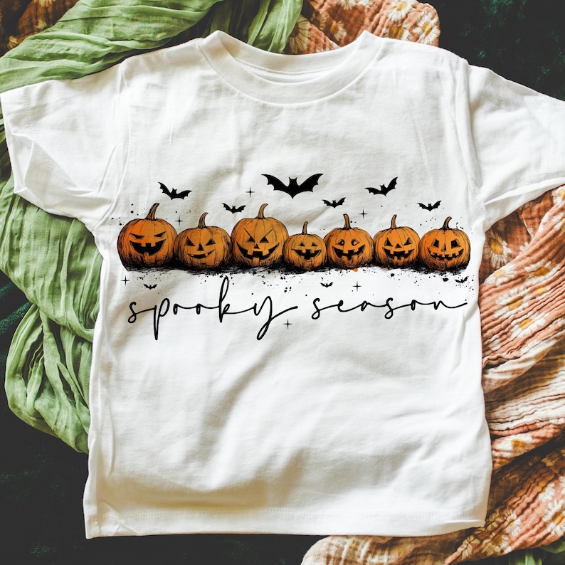 Op de afbeelding: Wit T-shirt met een rij uitgesneden oranje pompoenen met zwarte gezichten en vleermuizen. De tekst "spooky season" is in zwarte letters geschreven. Een Halloween-thema grafisch T-shirt.