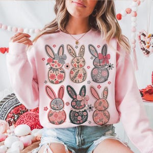 May include: Pink sweatshirt with a floral pattern of six bunnies wearing bows. The bunnies are in a row, with three on top and three on the bottom. The sweatshirt is a crew neck style with long sleeves.