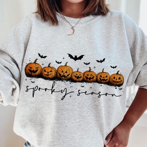 Op de afbeelding: Een lichtgrijs sweatshirt met een rij uitgesneden oranje pompoenen en zwarte vleermuizen. De tekst "spooky season" staat in zwart schrift onder de pompoenen. Een ketting met een maanvormige hanger is zichtbaar.