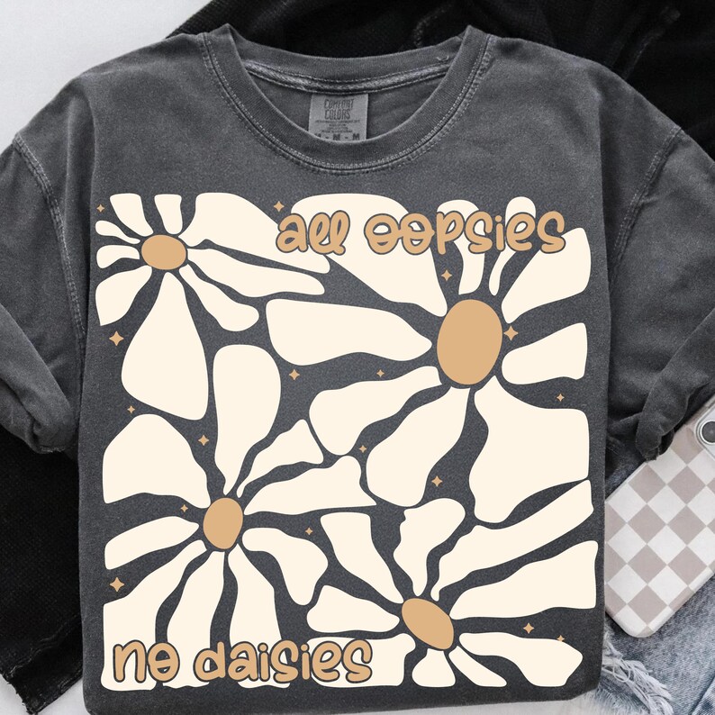 May include: A dark grey t-shirt with a graphic of stylised white daisies with brown centres. The text "all oopsies no daisies" is printed in brown on the shirt.