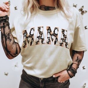 Pu&ograve; includere: T-shirt color crema con la scritta "MAMA" in un font in stile collegiale. Le lettere sono riempite con un motivo di fantasmi bianchi e fiori su sfondo nero, bordate di arancione. Un top casual a tema Halloween.