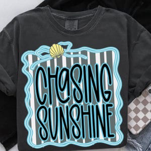 Puede incluir: Camiseta gris oscuro con las palabras "CHASING SUNSHINE" en un dise&ntilde;o azul y blanco. El dise&ntilde;o incluye una concha y un fondo a rayas. Una prenda de vestir informal.