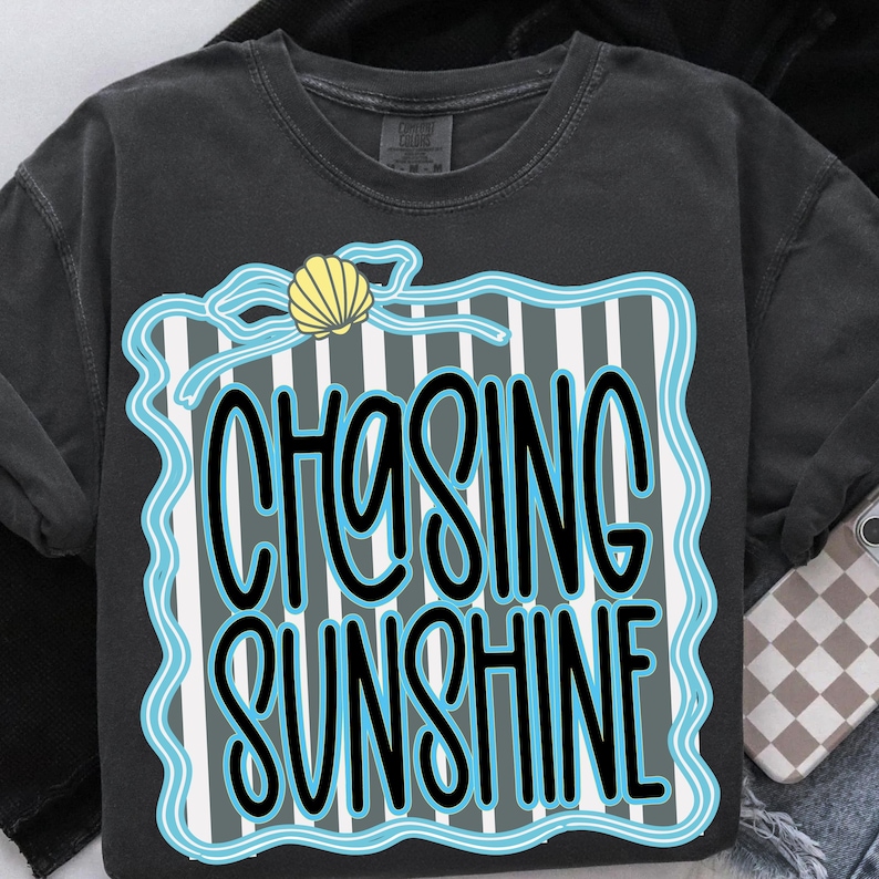 Puede incluir: Camiseta gris oscuro con las palabras "CHASING SUNSHINE" en un dise&ntilde;o azul y blanco. El dise&ntilde;o incluye una concha y un fondo a rayas. Una prenda de vestir informal.
