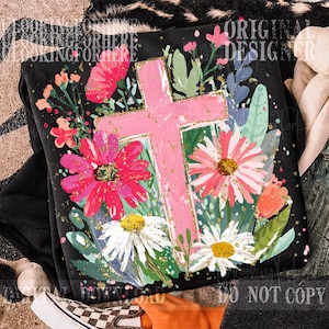 May include: Black sweatshirt featuring a pink cross design surrounded by colorful flowers and foliage. The cross is outlined in gold glitter, and the flowers include shades of pink, red, white, and green. The words "ORIGINAL DESIGNER" are printed on the top right.