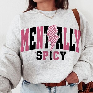 May include: A light gray sweatshirt with the words "MENTALLY SPICY" in black and pink block letters. A pink and white checkered lightning bolt graphic is centered above the word "SPICY". The sweatshirt has long sleeves.