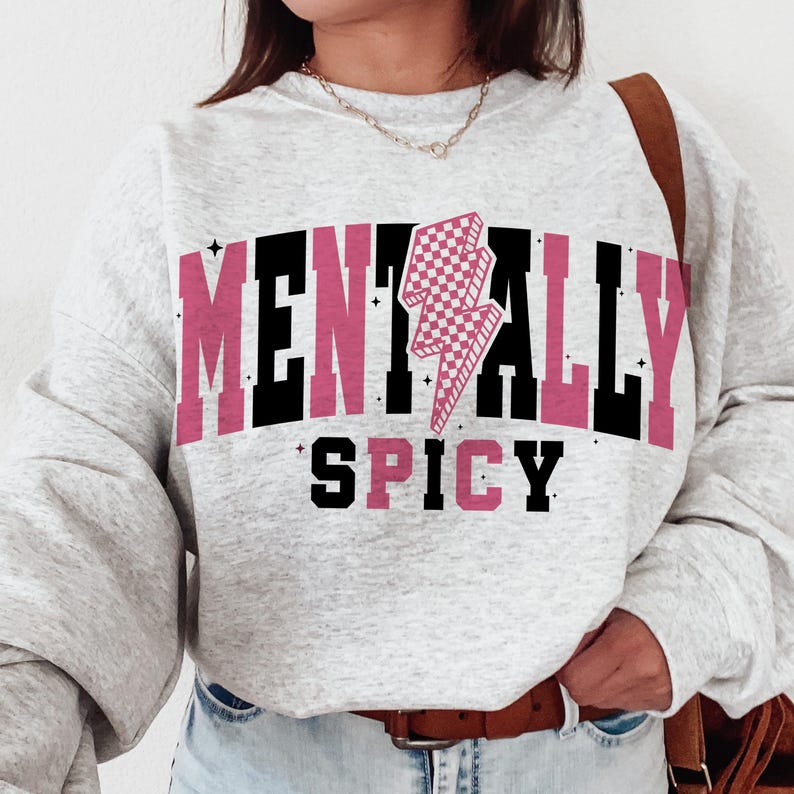 May include: A light gray sweatshirt with the words "MENTALLY SPICY" in black and pink block letters. A pink and white checkered lightning bolt graphic is centered above the word "SPICY". The sweatshirt has long sleeves.