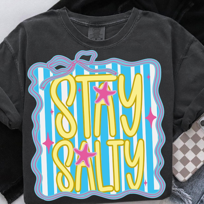 Puede incluir: Camiseta gris oscuro con la frase "STAY SALTY" en amarillo, sobre un fondo de rayas azules y blancas. El dise&ntilde;o incluye estrellas de mar rosas y un lazo rosa.