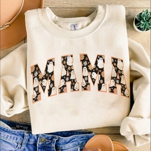 Pu&ograve; includere: Felpa color crema con la scritta "MAMA" in un font retr&ograve;. Le lettere sono riempite con un motivo floreale nero e illustrazioni di fantasmi bianchi. Un top elegante e confortevole per Halloween.