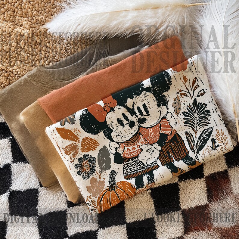 May include: Folded clothing items, including a tan, rust, and olive green sweatshirt, next to a patterned fabric featuring Mickey and Minnie Mouse. The fabric has a fall theme with pumpkins and leaves.