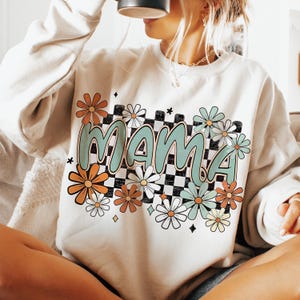 Puede incluir: Una sudadera beige claro con un estampado gr&aacute;fico de la palabra "mama" en un patr&oacute;n de cuadros verde y blanco con flores coloridas que rodean el texto.