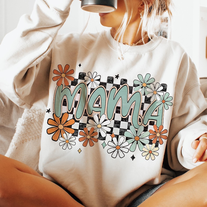 Puede incluir: Una sudadera beige claro con un estampado gr&aacute;fico de la palabra "mama" en un patr&oacute;n de cuadros verde y blanco con flores coloridas que rodean el texto.