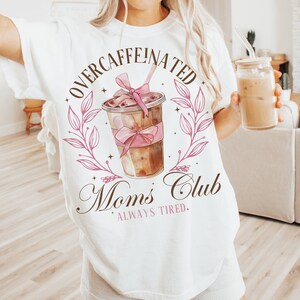 May include: White t-shirt with a brown and pink graphic of a coffee cup with a pink bow and the text "Overcaffeinated Moms Club Always Tired."