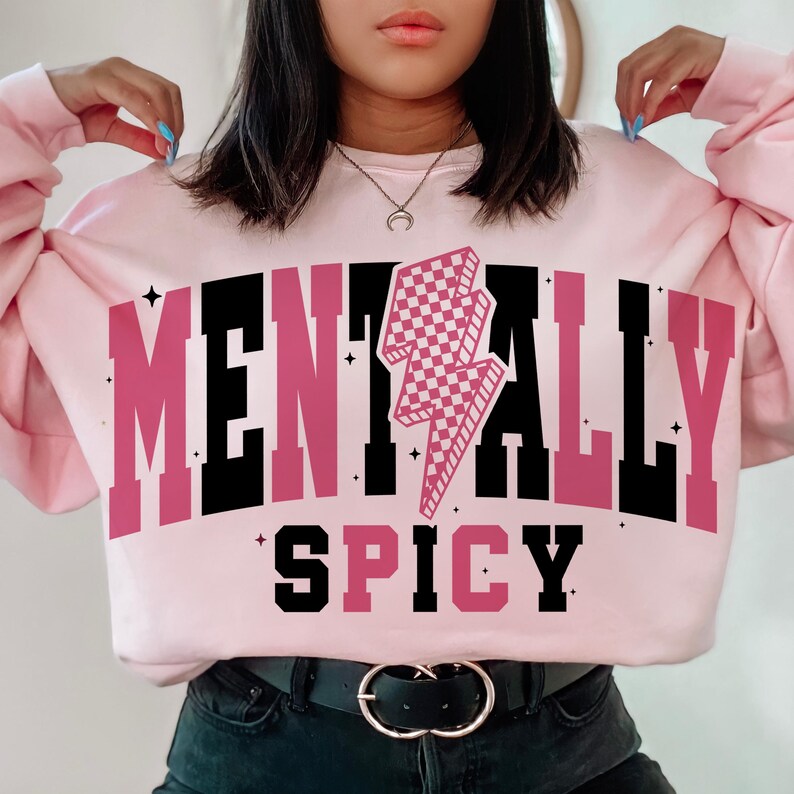 May include: A light pink sweatshirt with the words "MENTALLY SPICY" in a bold, two-tone font. A checkered pink lightning bolt graphic is centered above the word "SPICY". The sweatshirt has long sleeves and a crew neck.