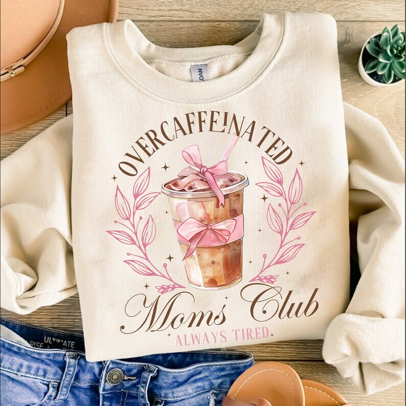 May include: A beige sweatshirt with a graphic of a coffee cup with a pink bow and the text "Overcaffeinated Moms Club Always Tired."