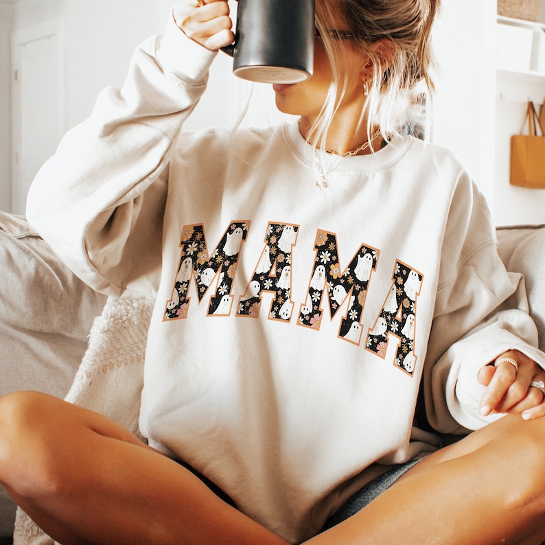 Pu&ograve; includere: Felpa beige con la scritta "MAMA" a grandi lettere. Le lettere sono riempite con un motivo floreale nero e illustrazioni di fantasmi bianchi. La felpa ha maniche lunghe e girocollo.