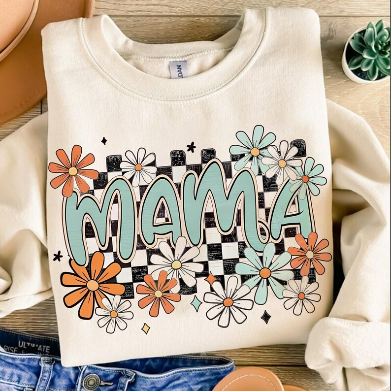Puede incluir: Una sudadera de color crema con un dise&ntilde;o gr&aacute;fico que presenta la palabra "MAMA" en color azul claro con un fondo a cuadros y flores de margarita de colores.