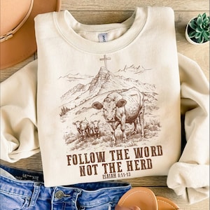 May include: A beige sweatshirt with a brown graphic of a cow and two calves in a mountain landscape with a cross on a peak. The text "FOLLOW THE WORD NOT THE HERD" and "ISAIAH 8:11-13" is printed below the graphic.