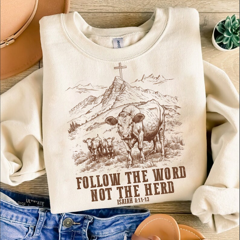 May include: A beige sweatshirt with a brown graphic of a cow and two calves in a mountain landscape with a cross on a peak. The text "FOLLOW THE WORD NOT THE HERD" and "ISAIAH 8:11-13" is printed below the graphic.