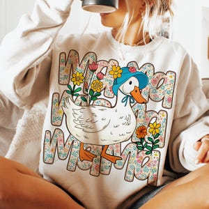 May include: A white sweatshirt with a cartoon duck wearing a blue hat and surrounded by colorful flowers. The text "Mama" is repeated multiple times around the duck.