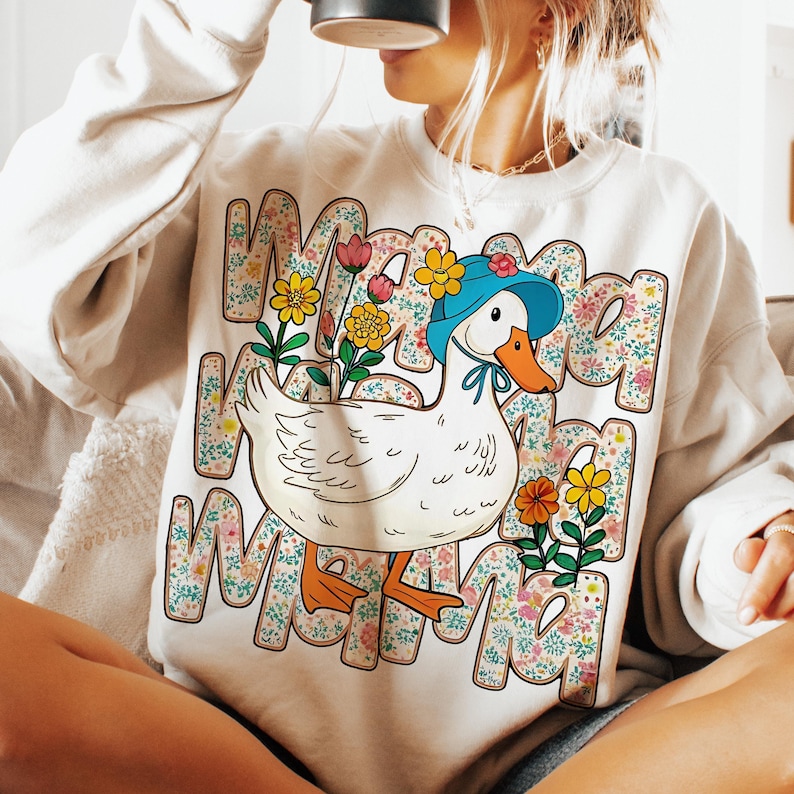 May include: A white sweatshirt with a cartoon duck wearing a blue hat and surrounded by colorful flowers. The text "Mama" is repeated multiple times around the duck.