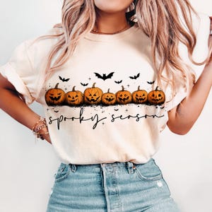 Op de afbeelding: Cr&egrave;mekleurig T-shirt met een rij uitgesneden oranje pompoenen met lachende gezichten en zwarte vleermuizen. De woorden "spooky season" staan eronder in zwarte letters. Een feestelijk Halloween-thema.