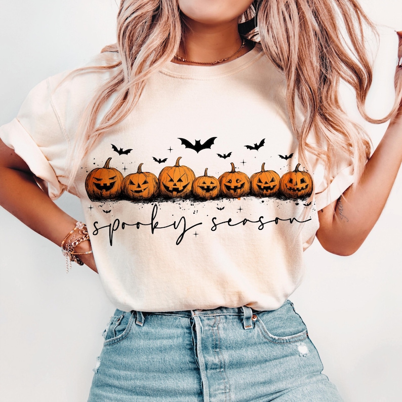 Op de afbeelding: Cr&egrave;mekleurig T-shirt met een rij uitgesneden oranje pompoenen met lachende gezichten en zwarte vleermuizen. De woorden "spooky season" staan eronder in zwarte letters. Een feestelijk Halloween-thema.