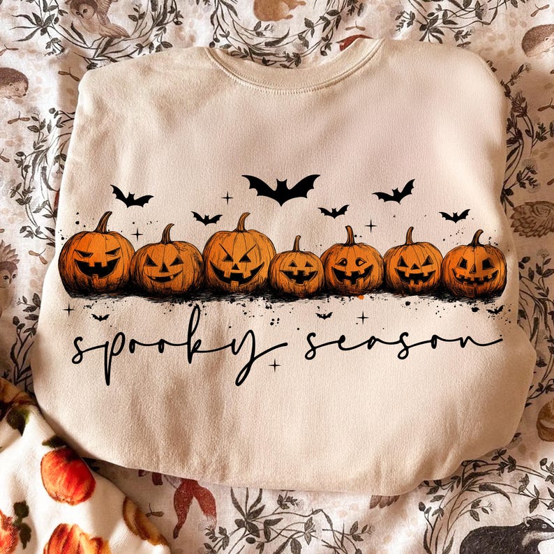 Op de afbeelding: Een beige sweatshirt met een rij uitgesneden oranje pompoenen, zwarte vleermuissilhouetten en de tekst "spooky season". De pompoenen hebben uitgesneden gezichten.