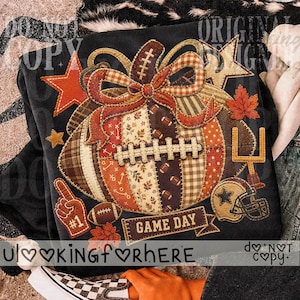May include: A black sweatshirt featuring a graphic of a football shaped like a pumpkin with a plaid and autumn-themed patchwork design. The graphic includes a bow, stars, a goal post, a football helmet, a foam finger, and the text GAME DAY.